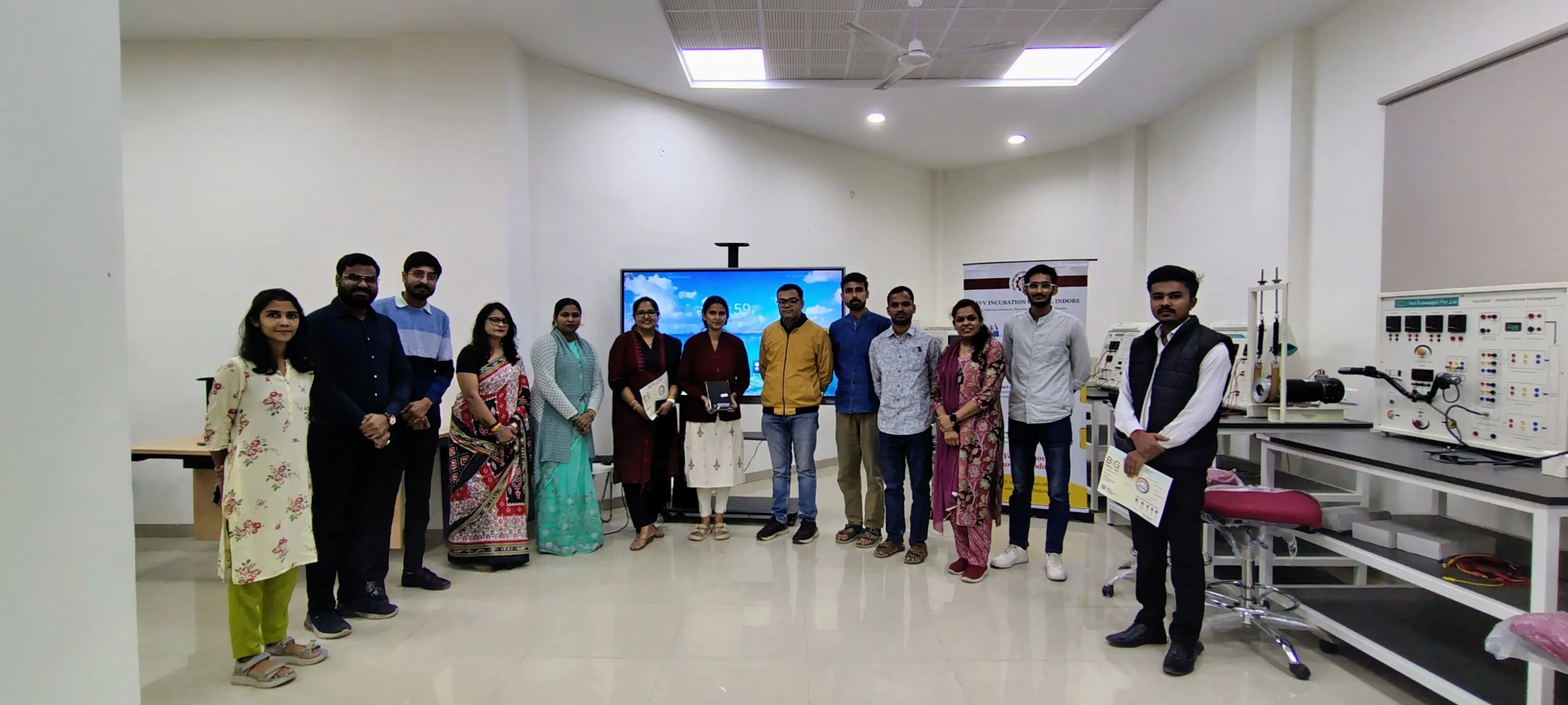 Group of people in a laboratory setting with equipment and a screen displaying content.